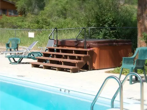 An outdoor hot tub with wooden steps sits beside a swimming pool.