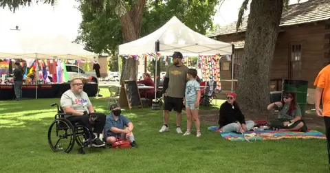 People gathered at an outdoor event on a grassy area near tents and a wooden building.