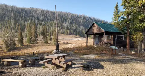 A wooden cabin sits in a clearing with a picnic table in the foreground and a forested mountain slope behind it.