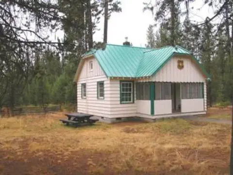 A small cabin with a green roof stands in a wooded area with a picnic table nearby.