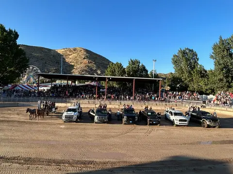 An outdoor arena with trucks and horses lined up before a large crowd of spectators.