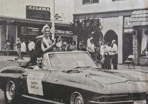 A queen rides in a convertible car down a street lined with buildings and spectators.