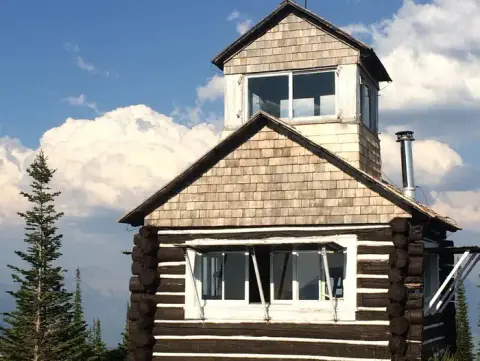 A rustic log cabin style lookout tower sits against a backdrop of mountains and cloudy sky.