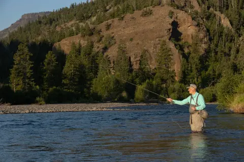 A person fly fishing in a river with mountains in the background.