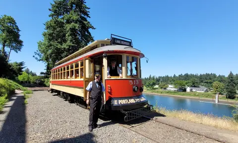 A vintage streetcar stopped on tracks next to a body of water with two operators visible.