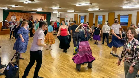 People dance inside a large hall with a wooden floor.