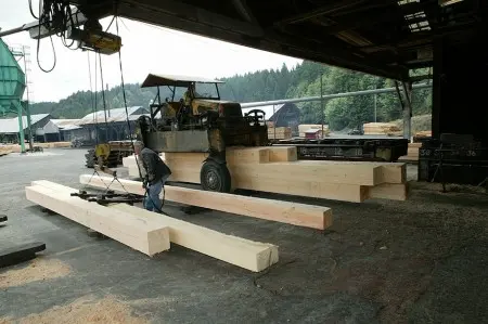A worker operates machinery handling large wooden beams at a timber mill.
