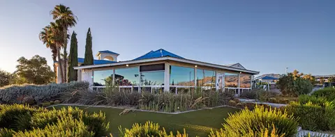 A single-story building with large windows and a blue roof is surrounded by desert landscaping and palm trees.