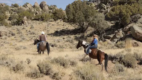 Two people ride horses through a dry, hilly landscape.