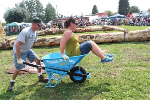 A man pushes a woman sitting in a blue wheelbarrow during an outdoor event.