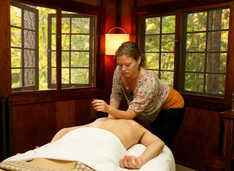 A massage therapist performs a massage in a wooden room with large windows overlooking trees.