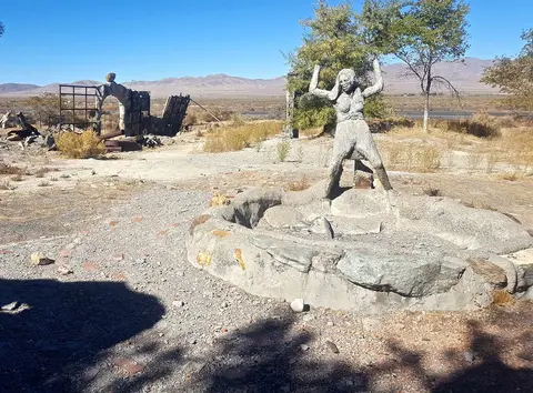 An outdoor art installation features a concrete statue of a human figure standing near ruins in a desert landscape.