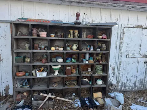 A large wooden shelving unit filled with various ceramic pottery items outdoors against a white wall.