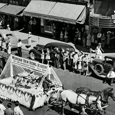 A historic black and white photo shows a parade with a horse-drawn float and spectators on a downtown street.
