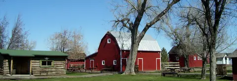 A red barn and rustic buildings stand under a clear blue sky on a grassy lot.