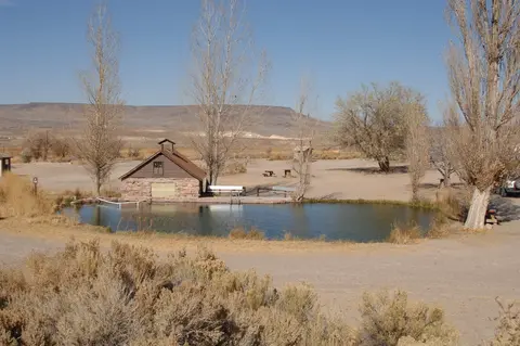 A stone and wood building stands beside a small pond in an arid landscape with sparse trees and distant hills.