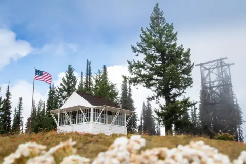 A small white building with an American flag and a tall wooden tower nearby outdoors.