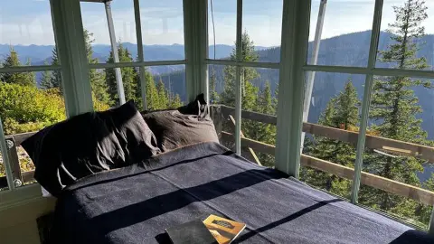 A bed inside a room with large windows overlooking a mountainous forested landscape.