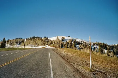 A road leading toward a snow-covered hill with trees under a blue sky.