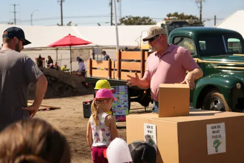A man in a pink shirt talks to children in construction hats near a green truck and a cardboard box.