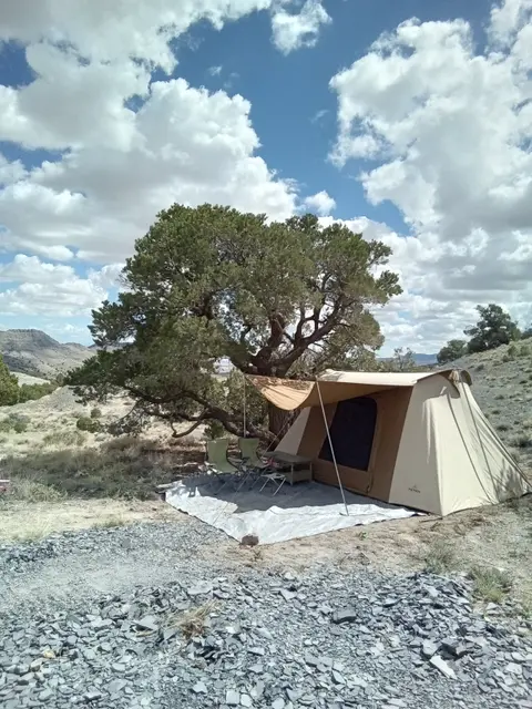 A tent and chairs are set up under a large tree in a rocky landscape with a cloudy sky.