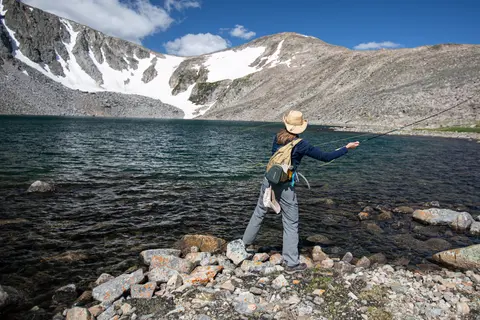 A person fly fishes in a mountain lake with snowy peaks in the background.