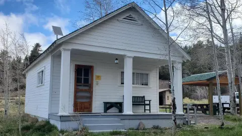 A small white building with a front porch and a solar panel on the roof stands among trees.