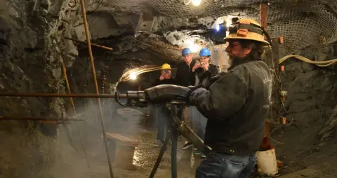 A man uses a pneumatic drill in an underground mine while others watch.