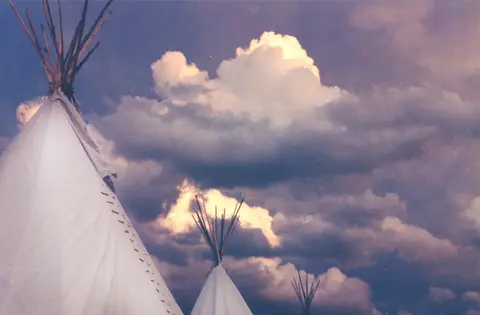 Three tipis stand against a dramatic sky filled with large clouds.