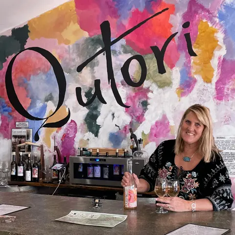 A woman holds a bottle of wine and glasses at a counter in front of a colorful painted wall.