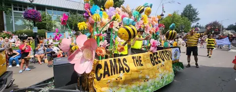 A decorated parade float featuring bees and flowers moves down a street with a crowd watching.