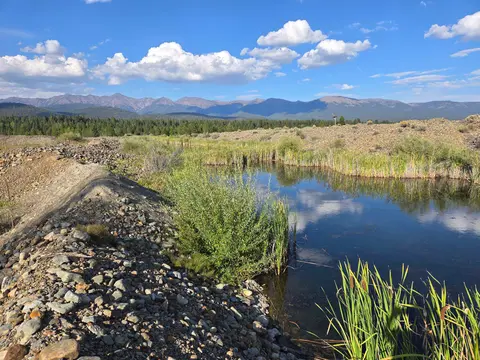 A calm pond reflects the blue sky and clouds, bordered by rocky shores and distant mountains.