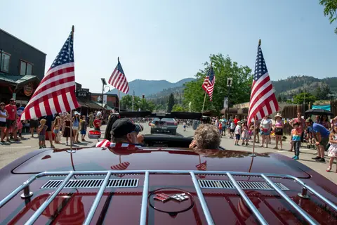 A vintage convertible with American flags drives down a street lined with spectators.