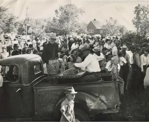Black and white historic photo showing people gathered around a pickup truck outdoors.
