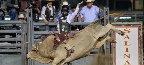 A cowboy rides a bucking bull at a rodeo while spectators watch from behind a fence.