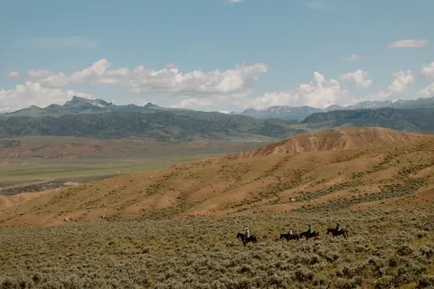 Four horseback riders traverse a rolling landscape beneath distant mountains.