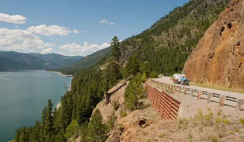 A truck with a camper drives along a mountain road next to a lake.