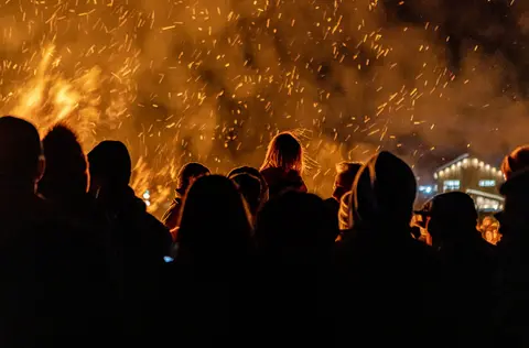 A crowd of people watch a large fire with glowing sparks filling the air.