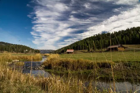 A scenic landscape featuring a winding stream through a grassy field with wooden structures and a forested hill under a dramatic cloudy sky.