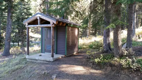 A small wooden outhouse with a covered porch sits in a forest.