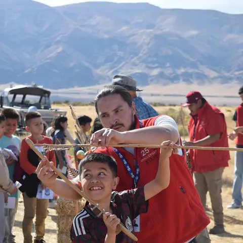 An adult helping a boy hold a long wooden stick outdoors with mountains in the background.