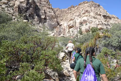 A group of people hiking on a trail in front of large rocky cliffs and bushes.