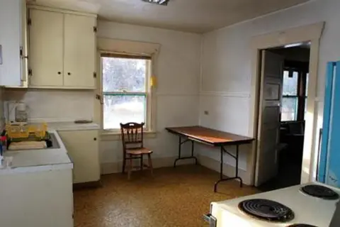 An interior view of a modest, older kitchen with cabinets, a table, and a chair.
