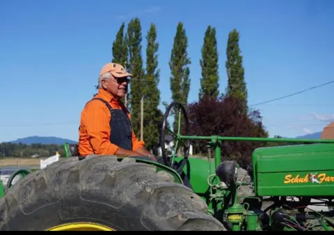 A man in an orange shirt and cap rides a green tractor.