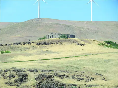 A stone structure on a grassy hillside with wind turbines in the background.