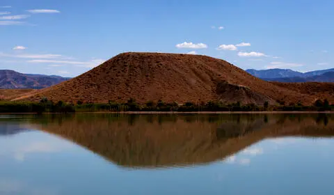 A brown hill is reflected in a calm body of water under a blue sky.