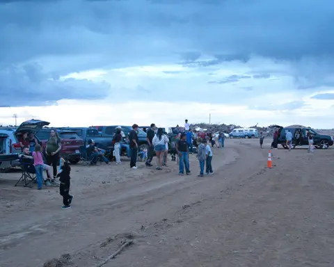 A crowd of people gathers near parked cars and trucks in a sandy area under a cloudy sky.