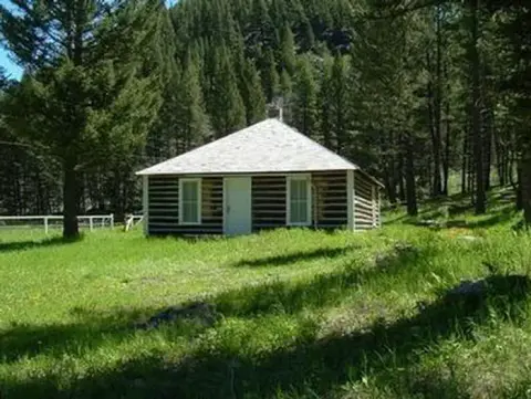 A log cabin sits in a grassy clearing surrounded by a dense forest.