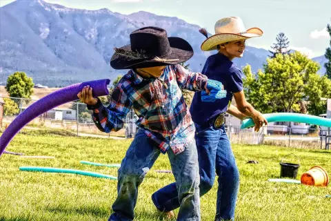 Two children wearing cowboy hats play in a grassy field with pool noodles.