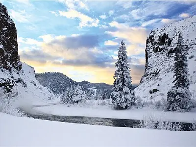A snowy landscape with a river flowing between rocky cliffs and pine trees.